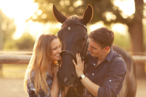 A young man and woman smile at each other while petting the nose of a brown horse that is standing between them.